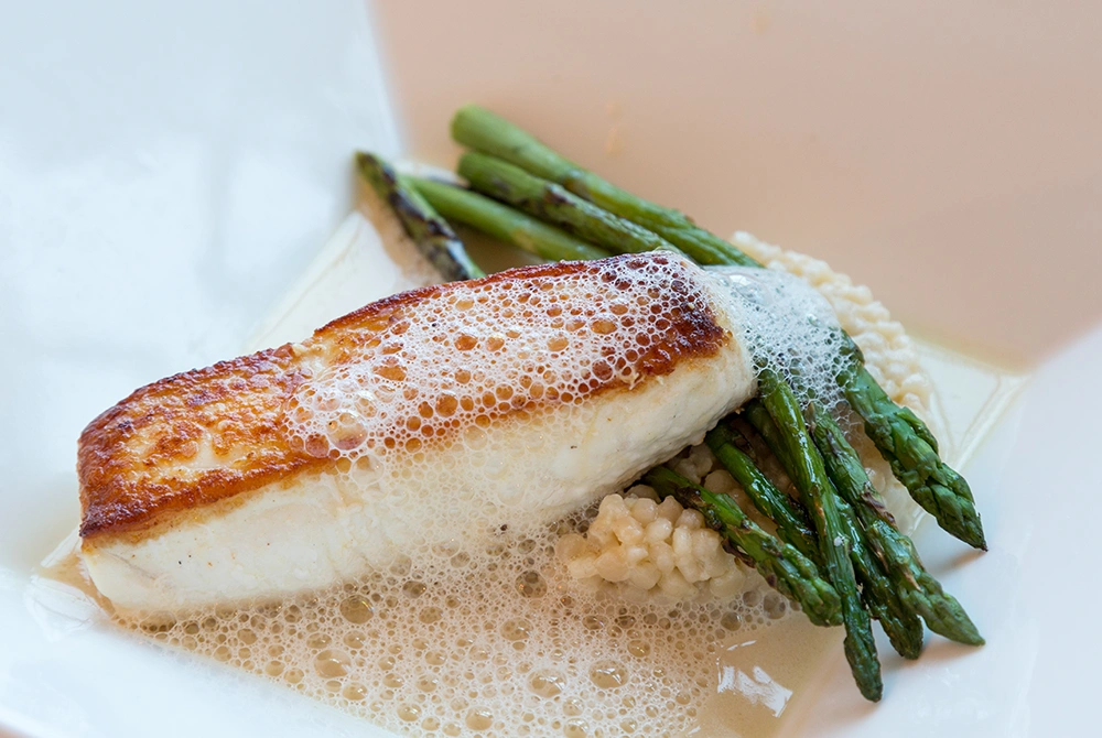 A seared fillet of fish sits on creamy risotto with grilled asparagus, topped with a light foam and elegantly presented on a white plate—an exquisite example of fine dining in Greenville ME.